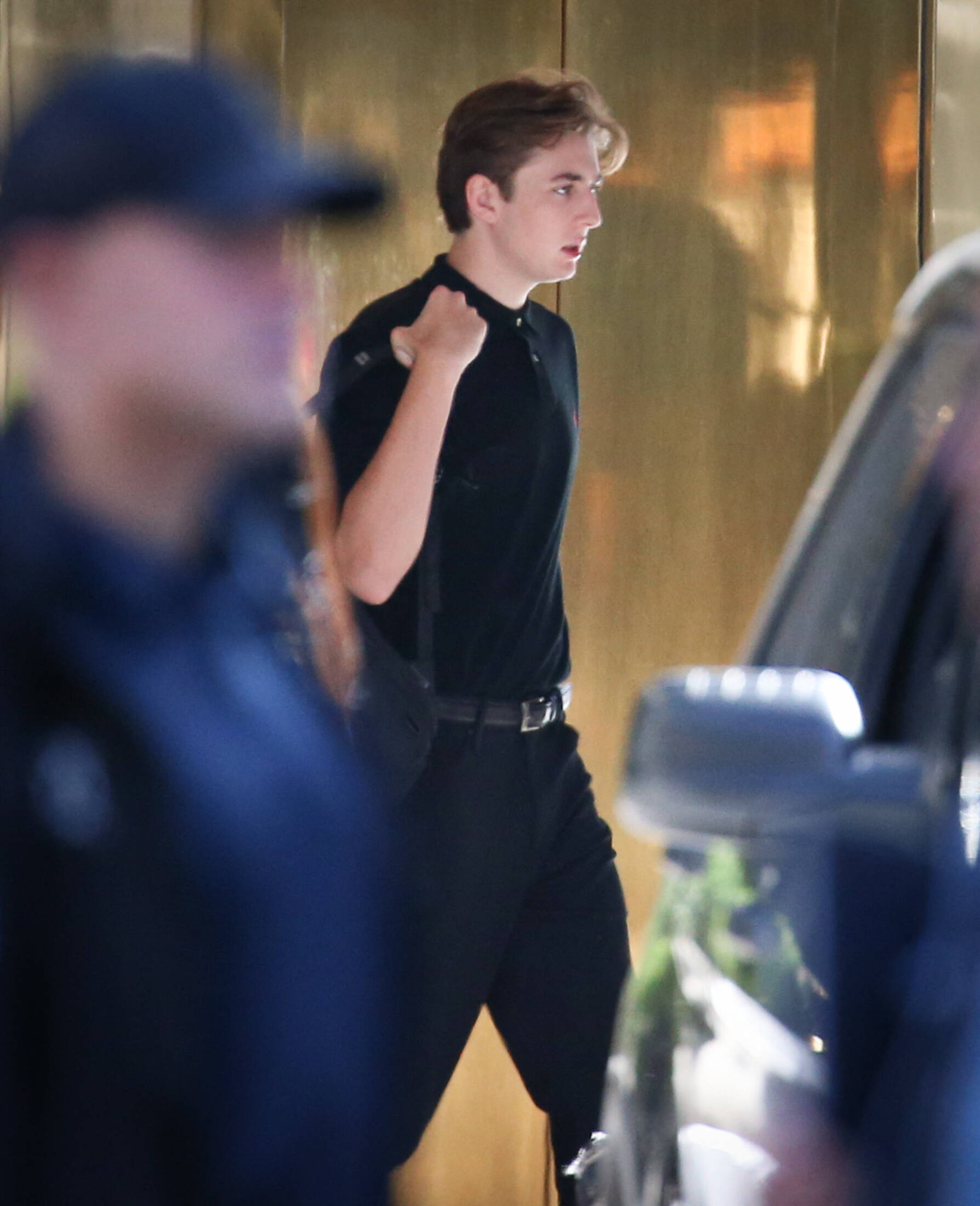 Barron Trump carries a bag over his shoulder while dressed in a black shirt and slacks, seen outside a building entrance.