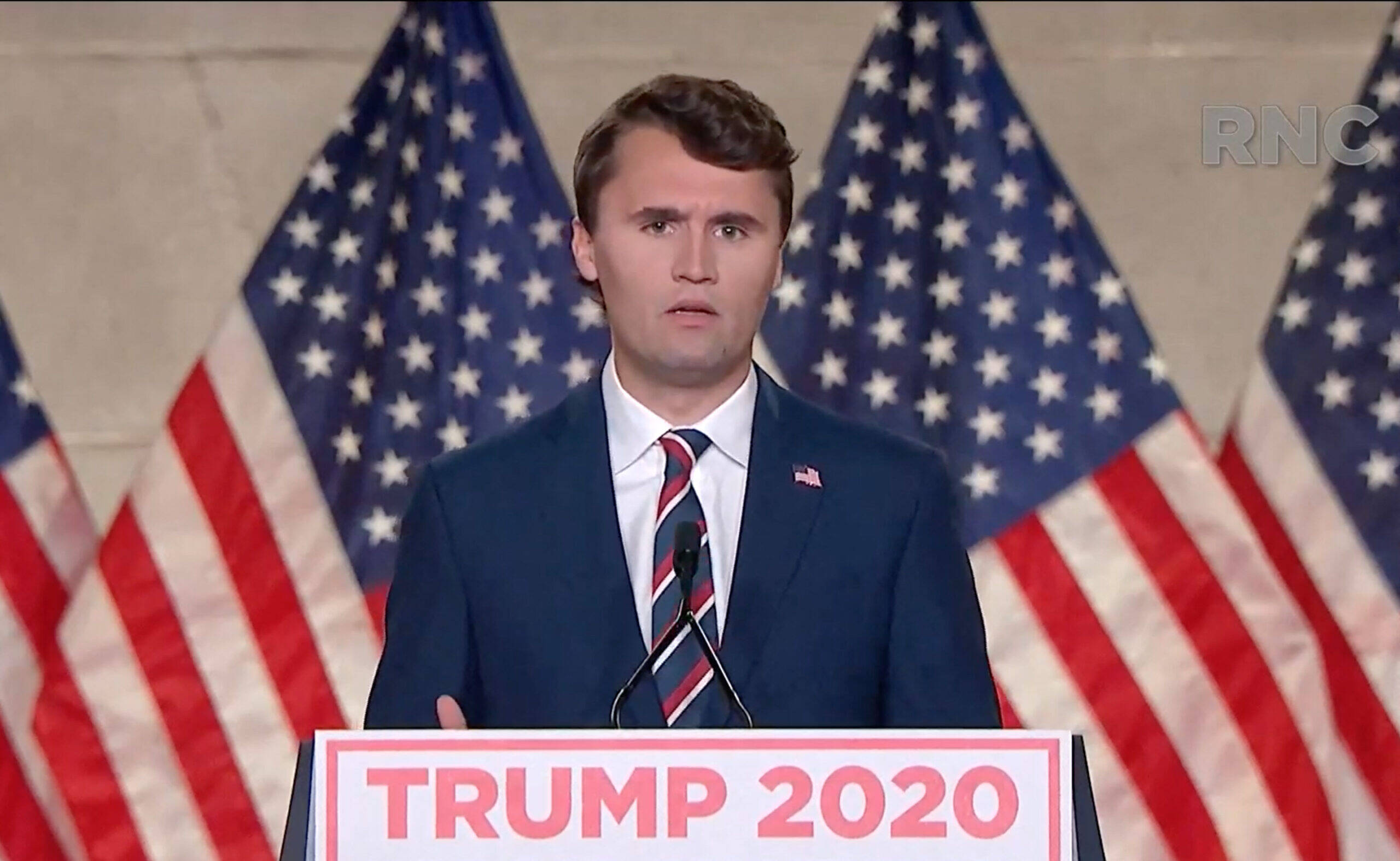 Charlie Kirk in a navy blue suit and striped tie speaks at a podium with “Trump 2020” signage, standing in front of American flags.